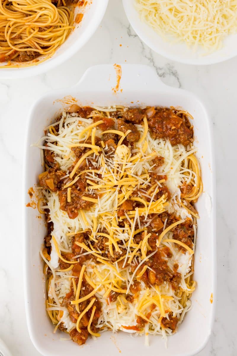 white baking dish with spaghetti, grated mozzarella and a scoops of meat sauce and shredded cheese, bowls of spaghetti and mozzarella in the background.