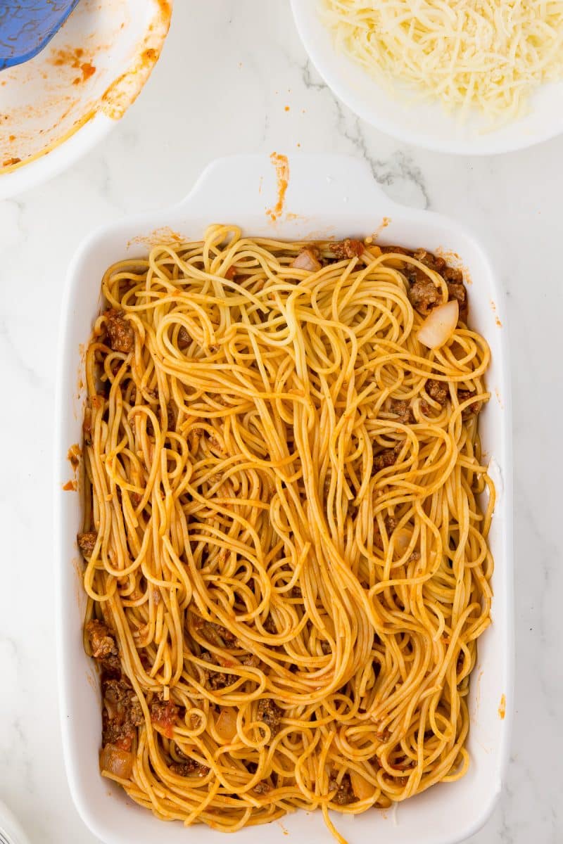white baking dish with spaghetti, grated mozzarella and a scoops of meat sauce, bowls of spaghetti and mozzarella in the background.