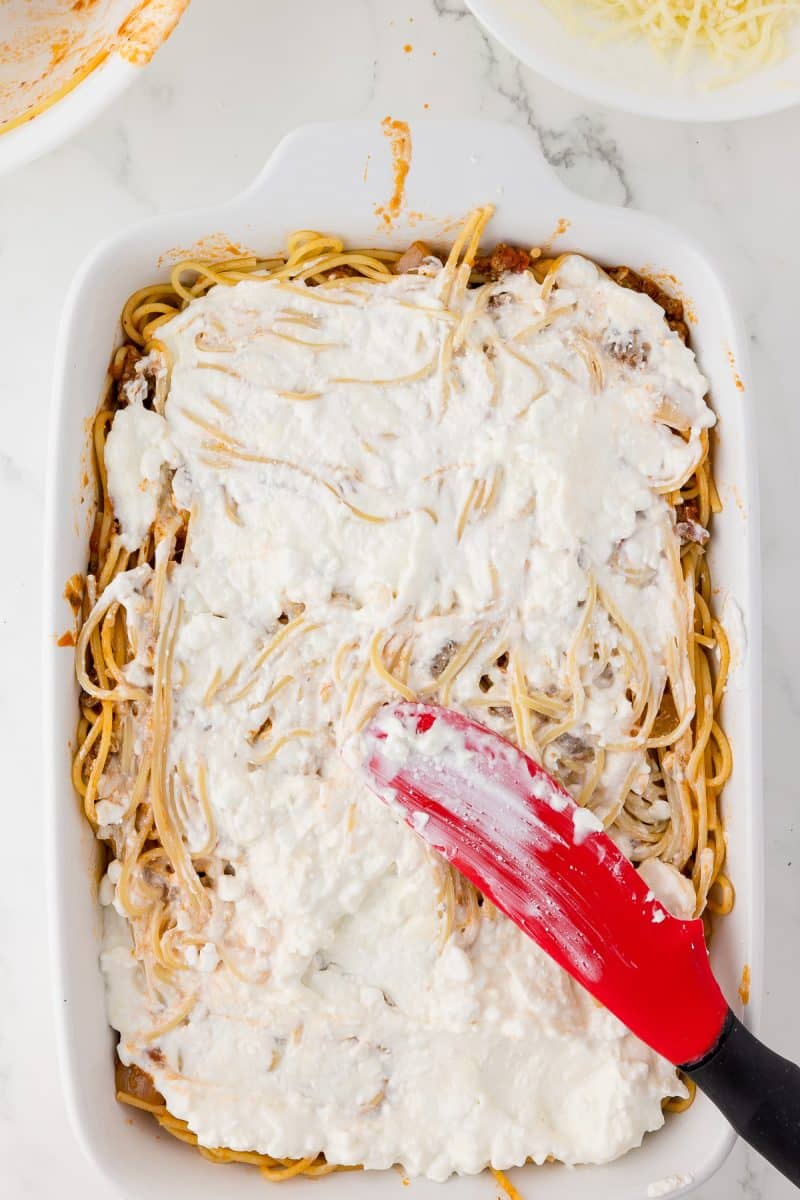 white baking dish with spaghetti, topped with cream cheese mixture, bowls of spaghetti and mozzarella in the background.