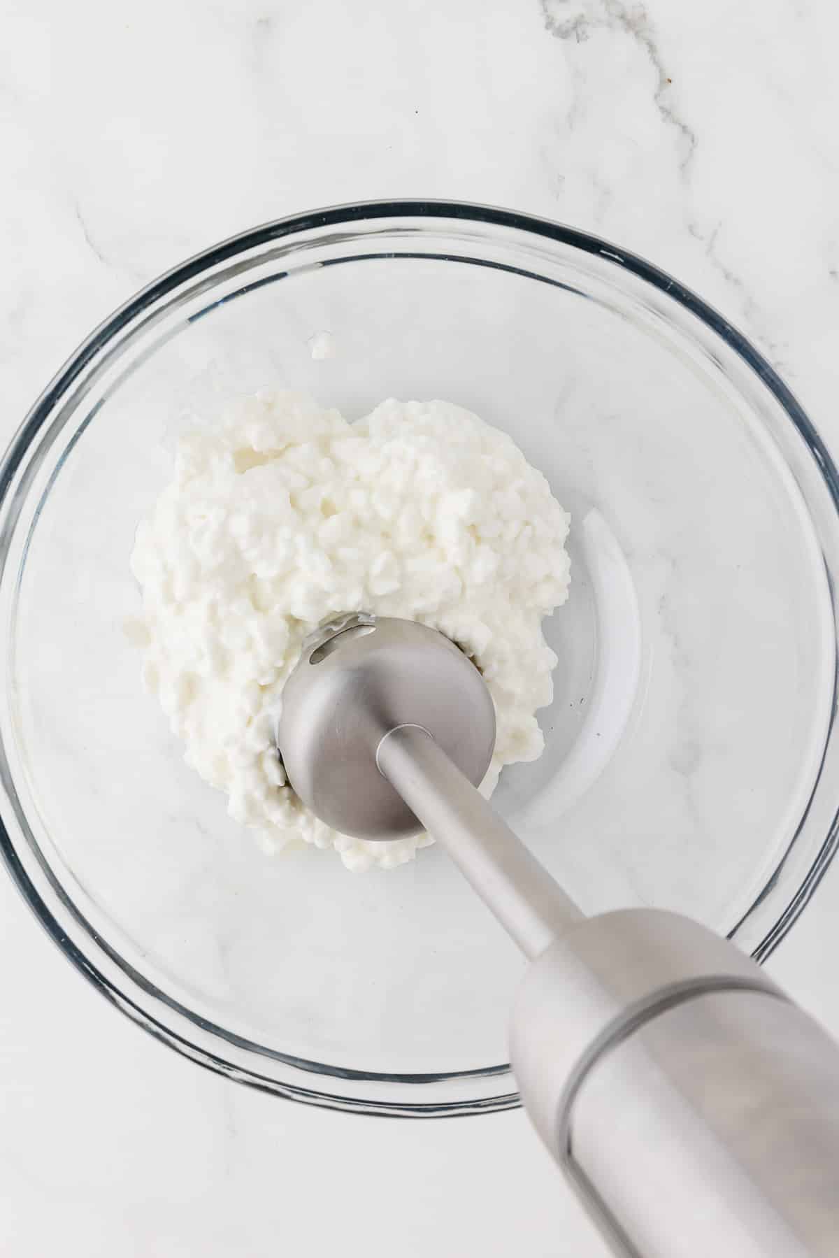 cottage cheese in a glass bowl being ground up by an immersion blender.