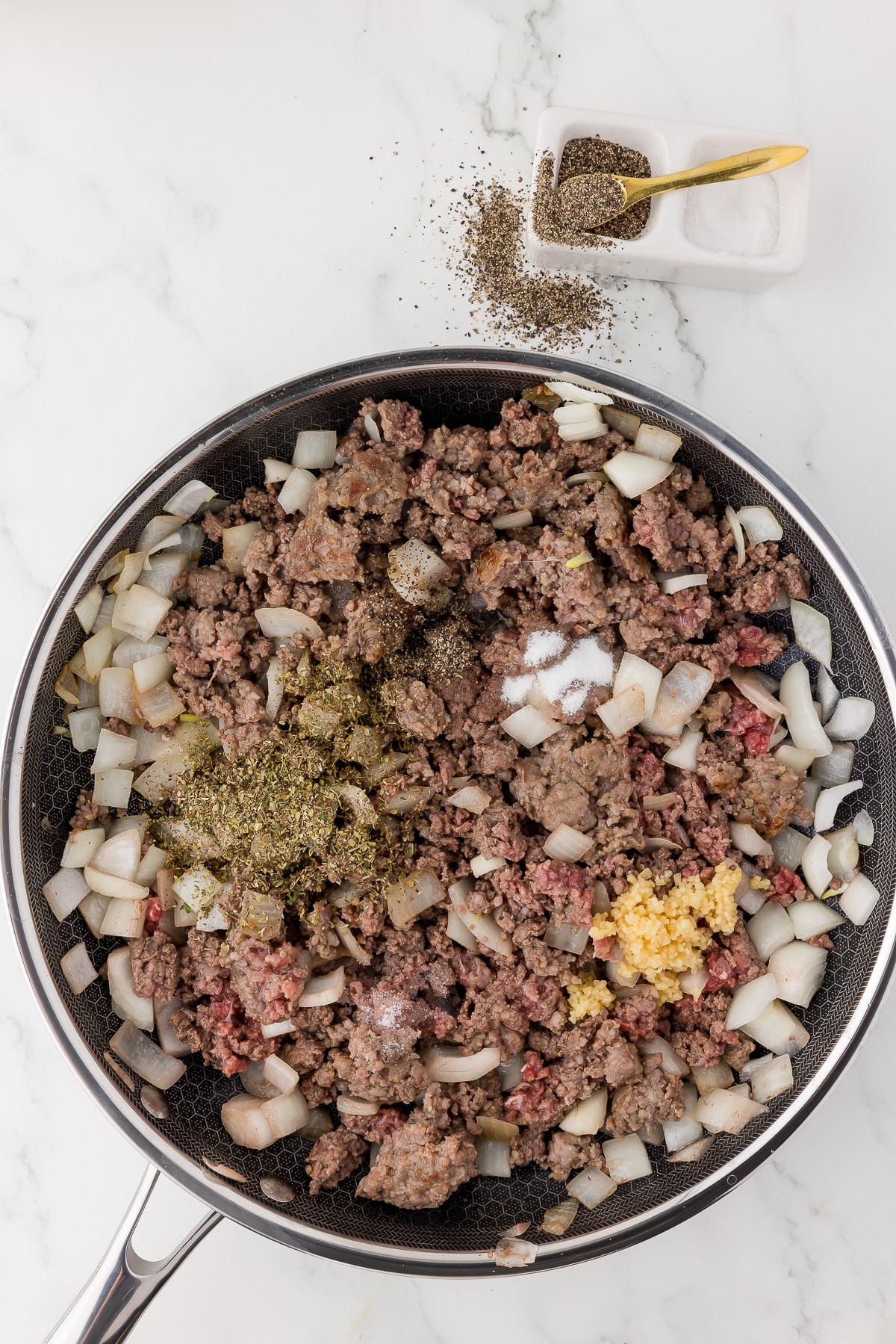 Ground meat, ground sausage, chopped onions, and spices in a Gordon Ramsay hexclad pan on a white marble countertop with salt and pepper in the background.