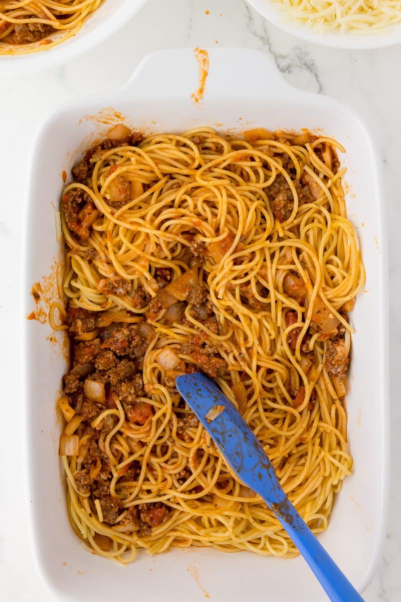 white baking dish with spaghetti, grated mozzarella and meat sauce