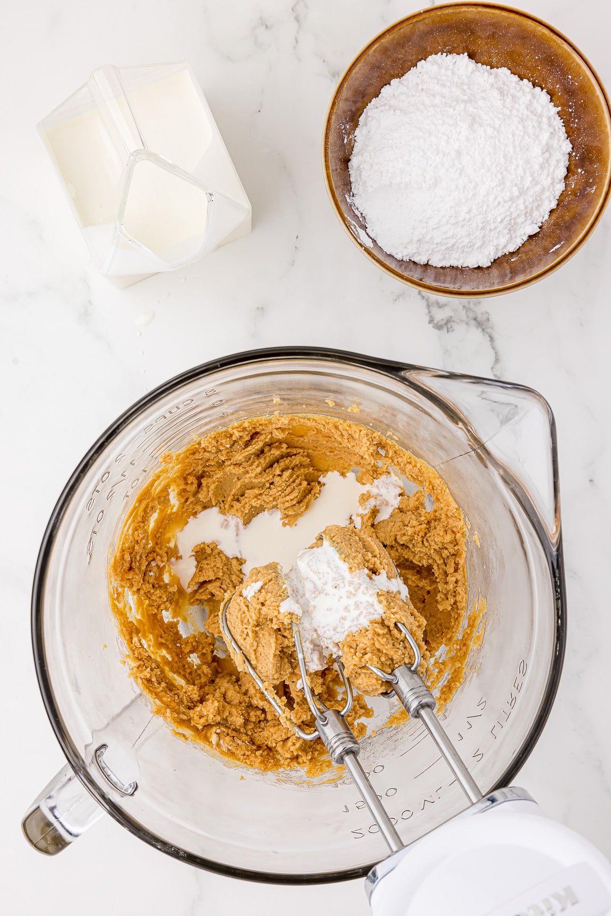 peanut butter filling in a glass batter bowl with kitchen aid mixer on a white countertop with cream and powdered in a bowl