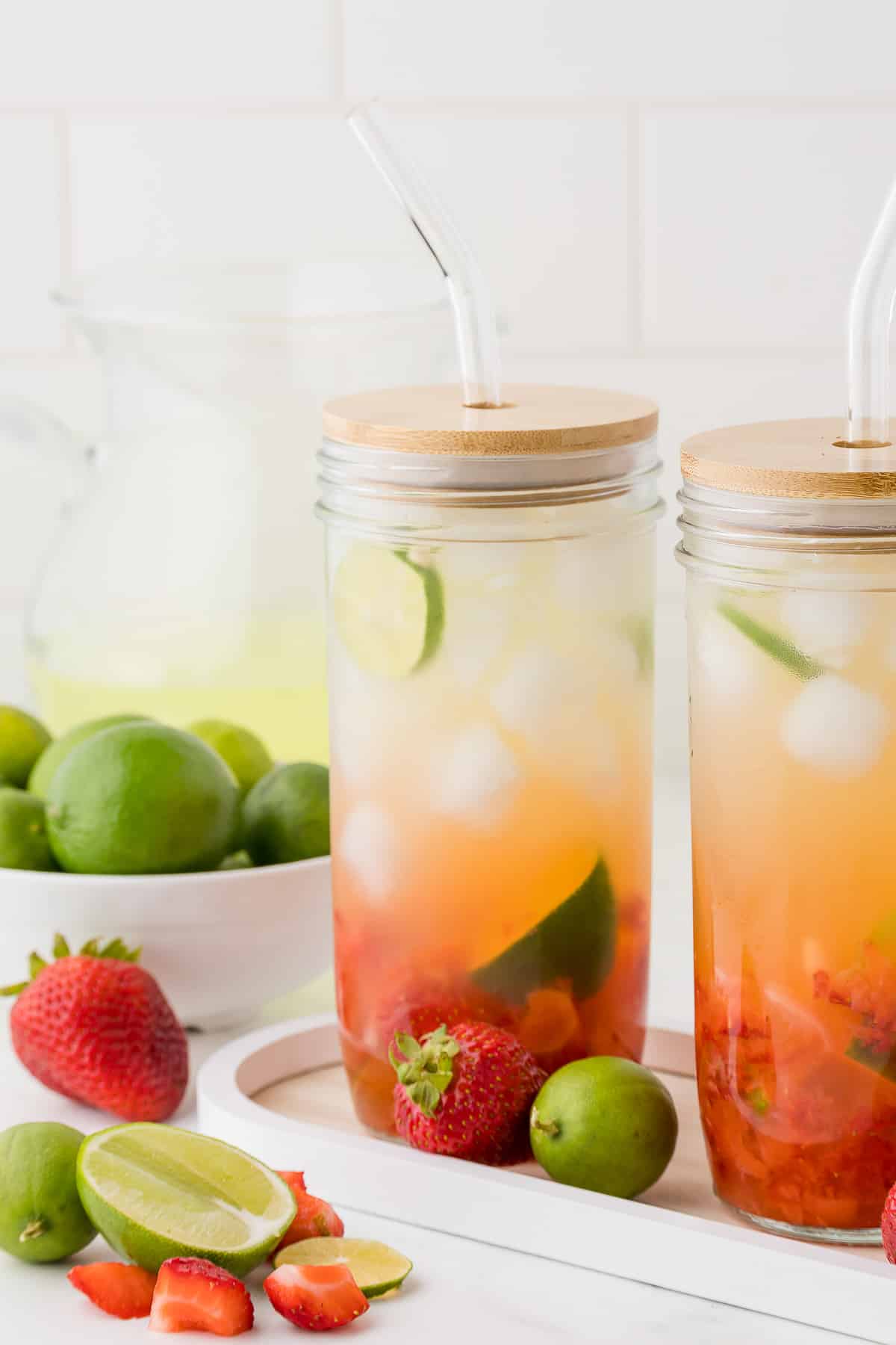 Two tall clear glass jars full of strawberry limeade with a bowl of limes in the background