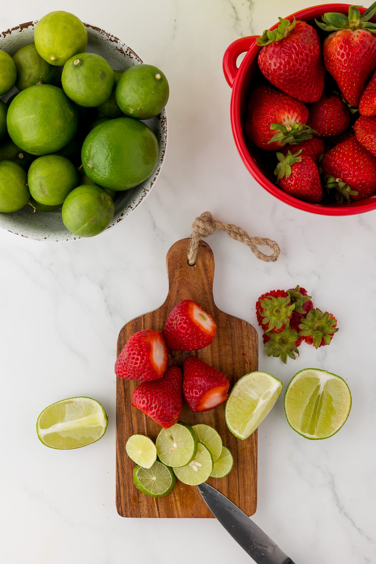 strawberries and limes on a wooden cutting board next to a large bowl of limes and a red bowl of strawberries