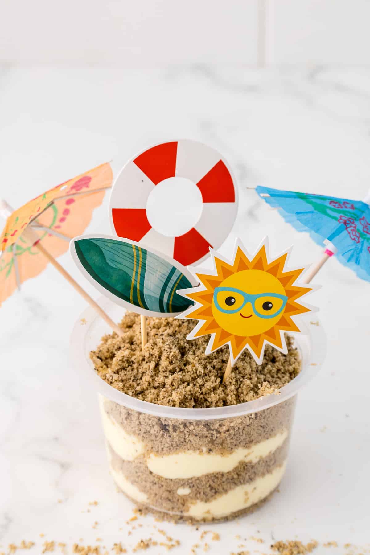 A clear plastic cup filled with pudding and sand made out of crumbled cookies with fun beach graphics.
