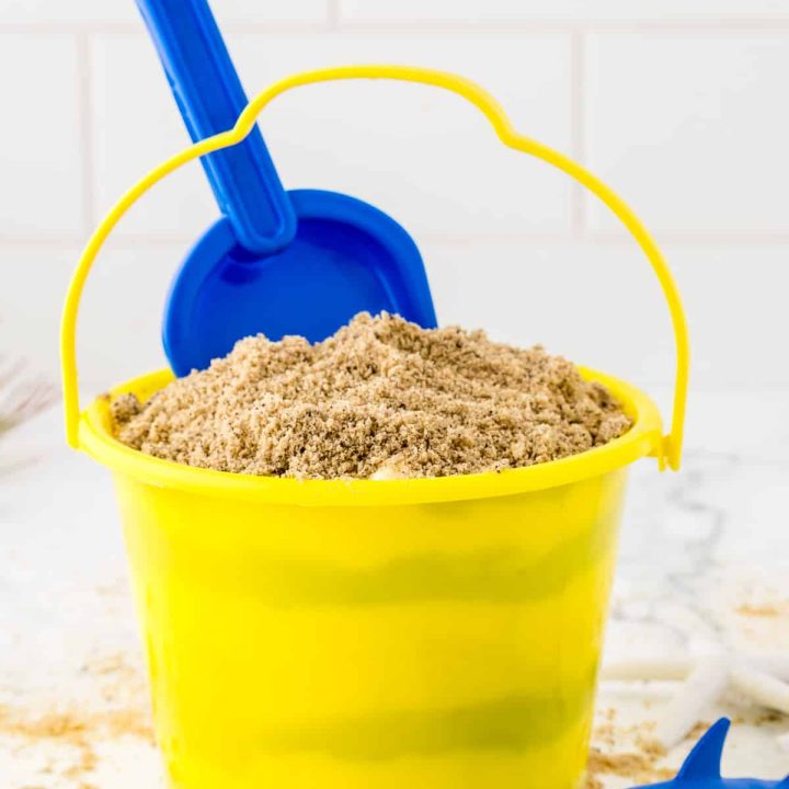 A yellow sand pail filled with pudding and topped with crumbled edible sand made from cookies including a blue sand shovel and plastic sharks