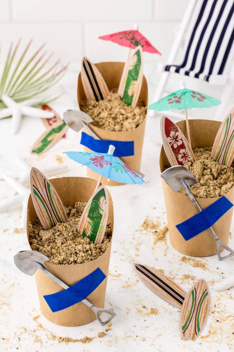 Three beach themed sand pudding dessert cups with paper umbrellas and surfboards.