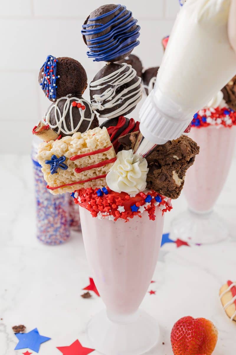 Strawberry milkshake with decorated assorted cookies coming out of the top, wilton sprinkes in the background additional whipping cream being added.