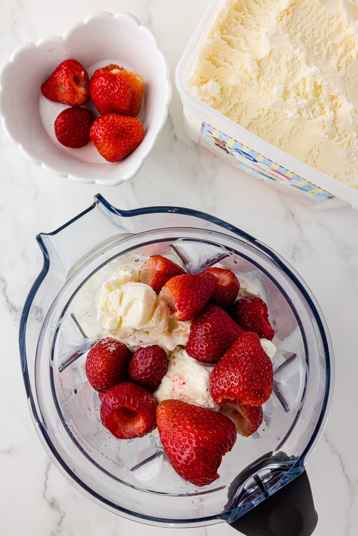 inside of a blender filled with vanilla ice cream and fresh strawberries, a bowl of strawberries and a carton of vanilla ice cream in the background on a white marble countertop.