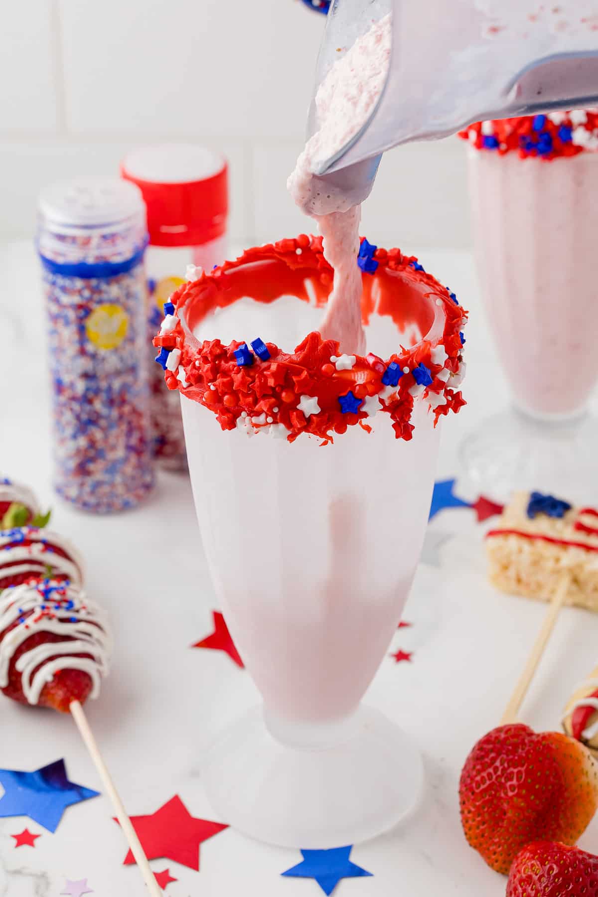 strawberry milkshake being poured into a frosted shake glass that is decorated with red frosting on the rim and red white and blue sprinkle stars.