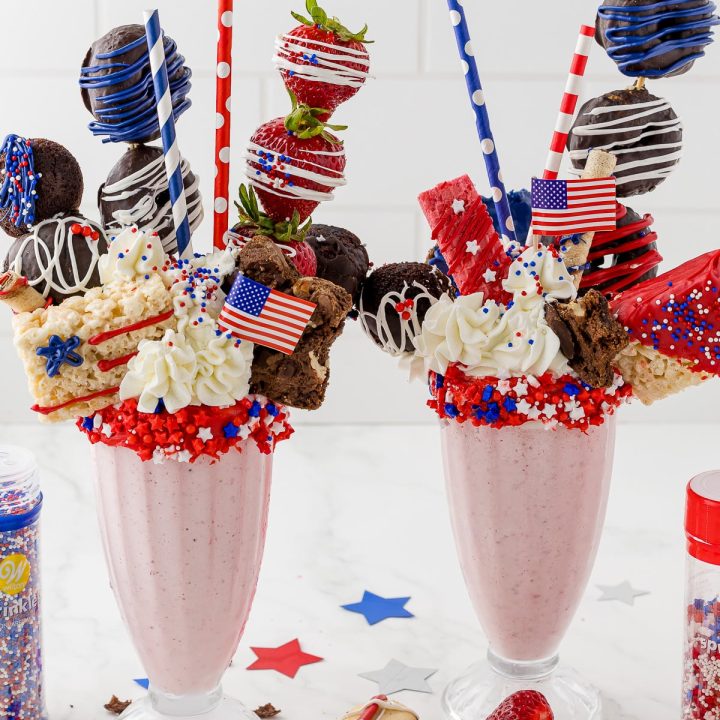 two strawberry milkshakes with patriotic decorated cookies stacked on top, red white and blue straws and wilton sprinkles.