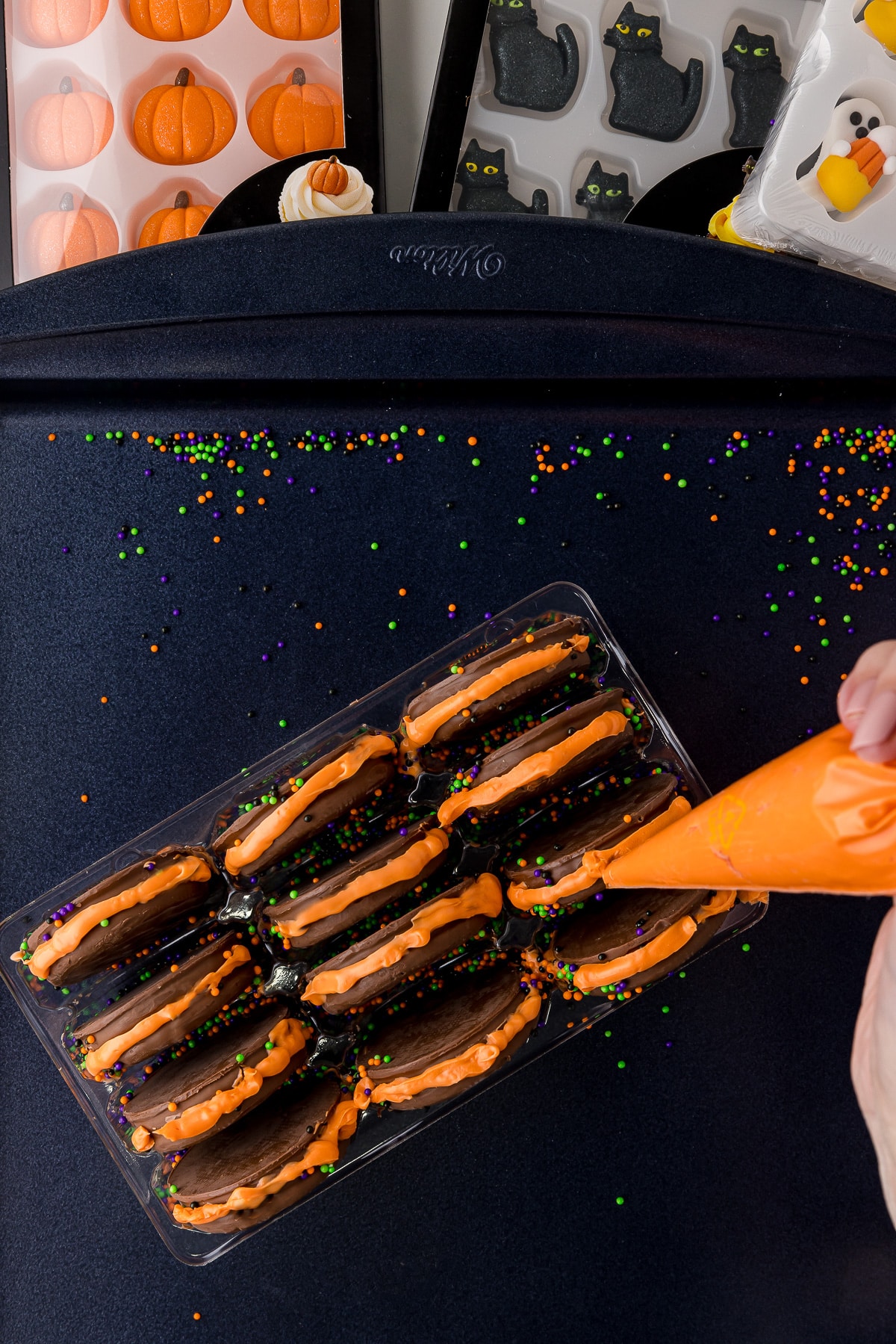 Orange melted candy being piped onto the sides of oreos in a dark cookie sheet.