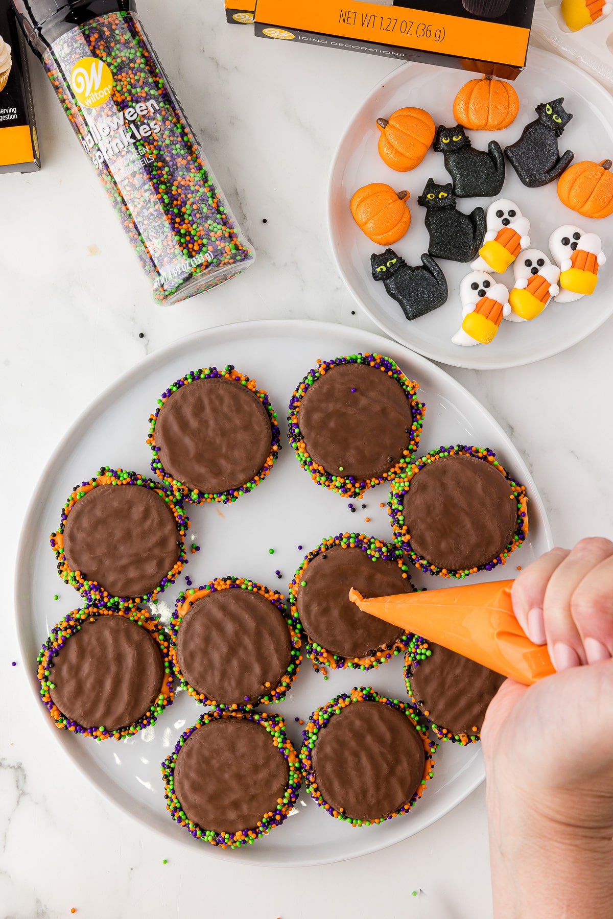 Orange melted chocolate dot on a fudge covered oreos on a white plate with halloween decorations and sprinkles on the counter. 