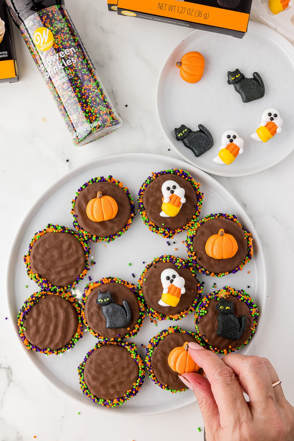 Wilton icing candy on fudge covered oreos on a white plate with halloween decorations and sprinkles on the counter. 