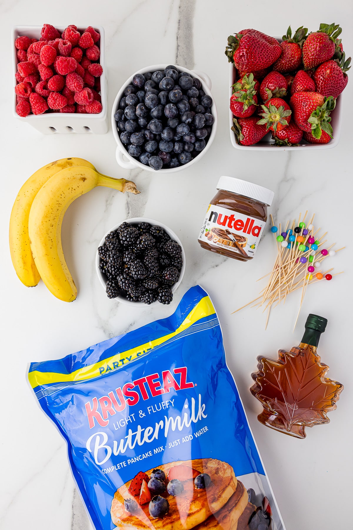 ingredients for mini pancakes including raspberries, blueberries, strawberries, bananas, blackberries, nutella, mable syrup, and Krusteaz buttermilk pancake mix