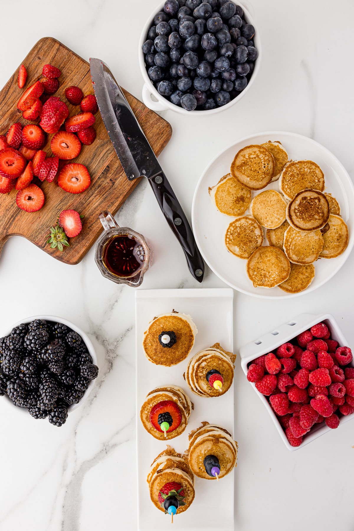overhead photos of a cutting board with sliced strawberries, a bowl each of blackberries, blueberries, and strawberries, with a serving plate of mini pancakes