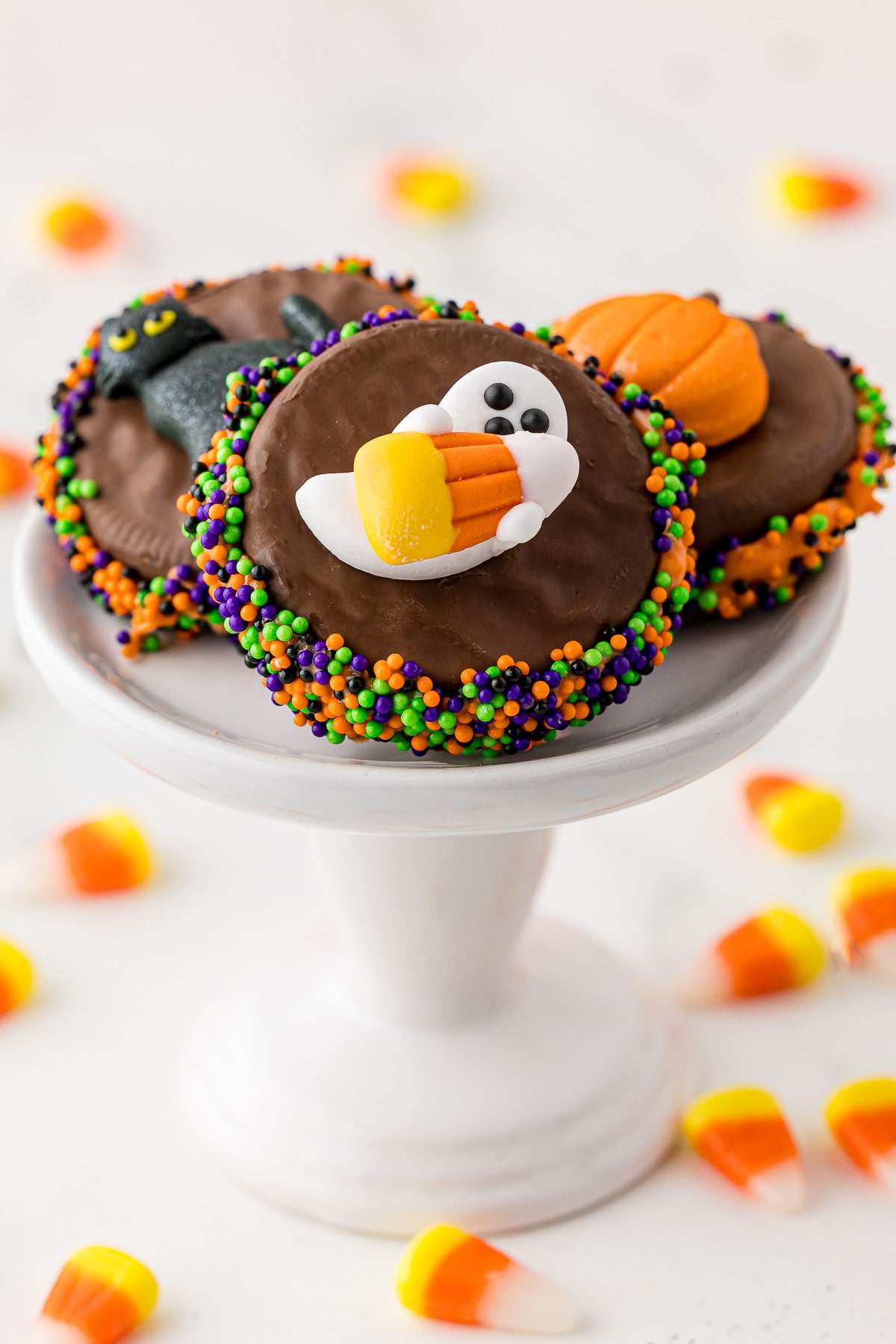 Chocolate covered Oreo cookie with orange, purple, green, and black Wilton sprinkles on the edges and a white candy ghost holding a candy acorn. Three like cookies sitting on a white cupcake stand with candy corn on the white marlbe countertop. 