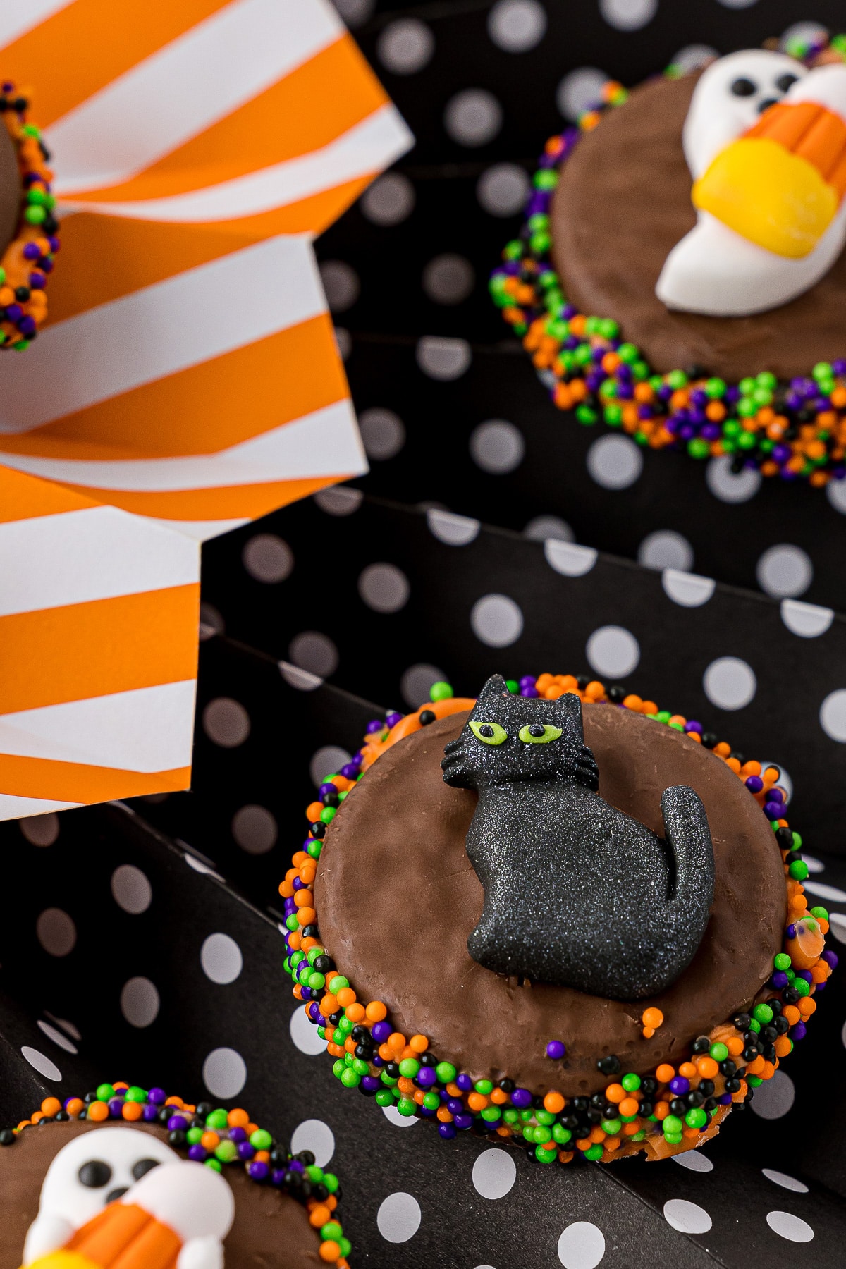 Black cat icing decoration on an oreo with orange, purple, green and black sprinkles sitting in a black and white polka dot fan and orange and white striped fan. 
