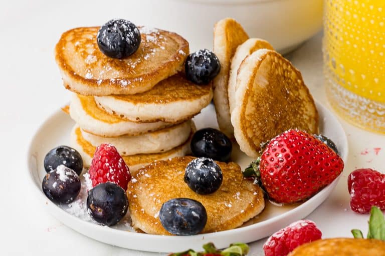 stack of mini pancakes with fruit on a white plate with a glass of orange juice in the background