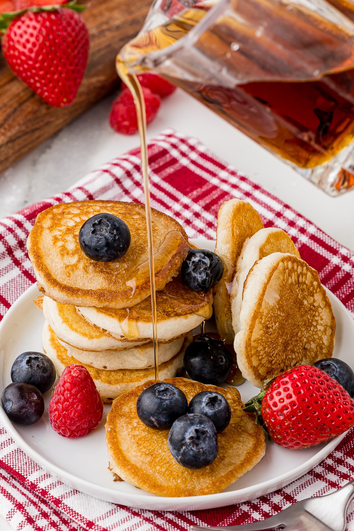 syrup being poured over mini pancakes stacked on a white plate with blueberries and strawberries