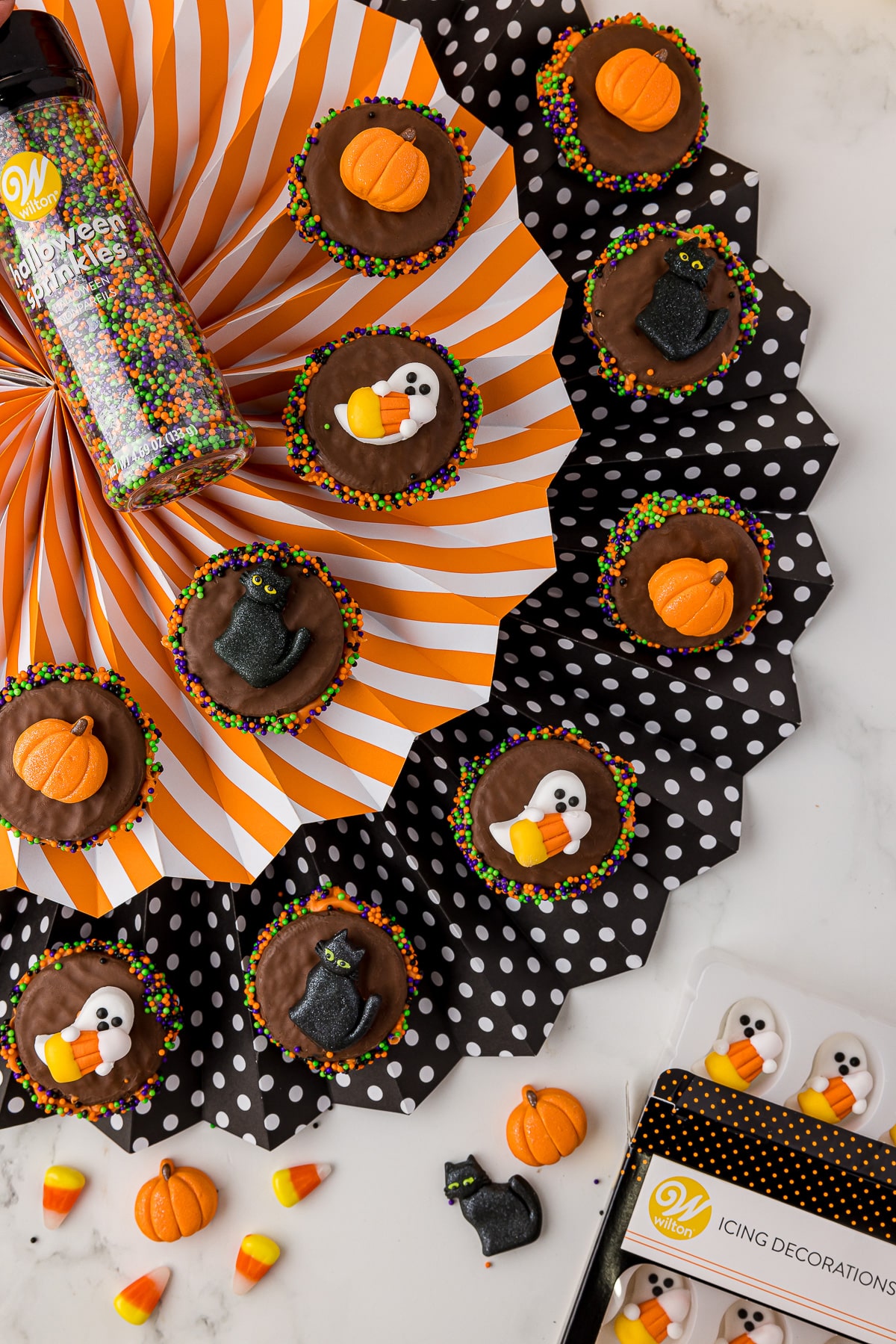 Decorated Halloween oreos swith orange, purple, green and black sprinkles sitting in a black and white polka dot fan and orange and white striped fan. 