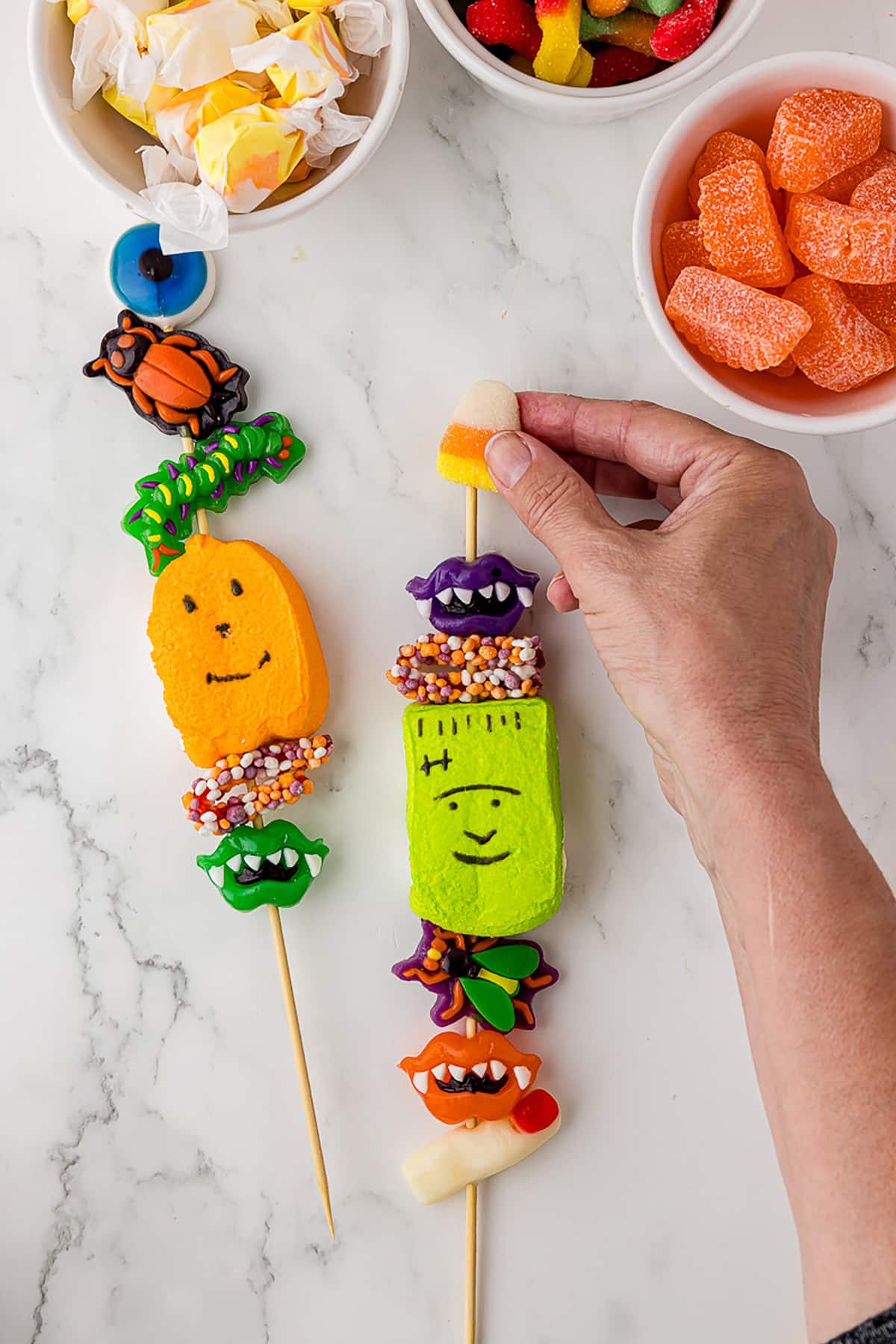 a hand putting a jelly candy corn on top of a skewer of halloween candy with an additional kabo, and three bowls of candy in the background