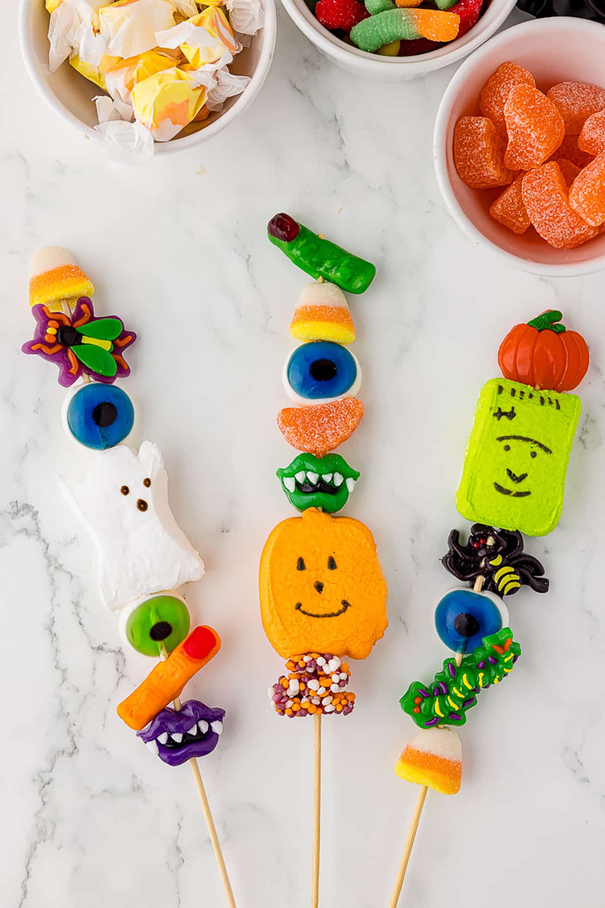 3 halloween candy kabobs with 3 bowls of gummy candy in the background