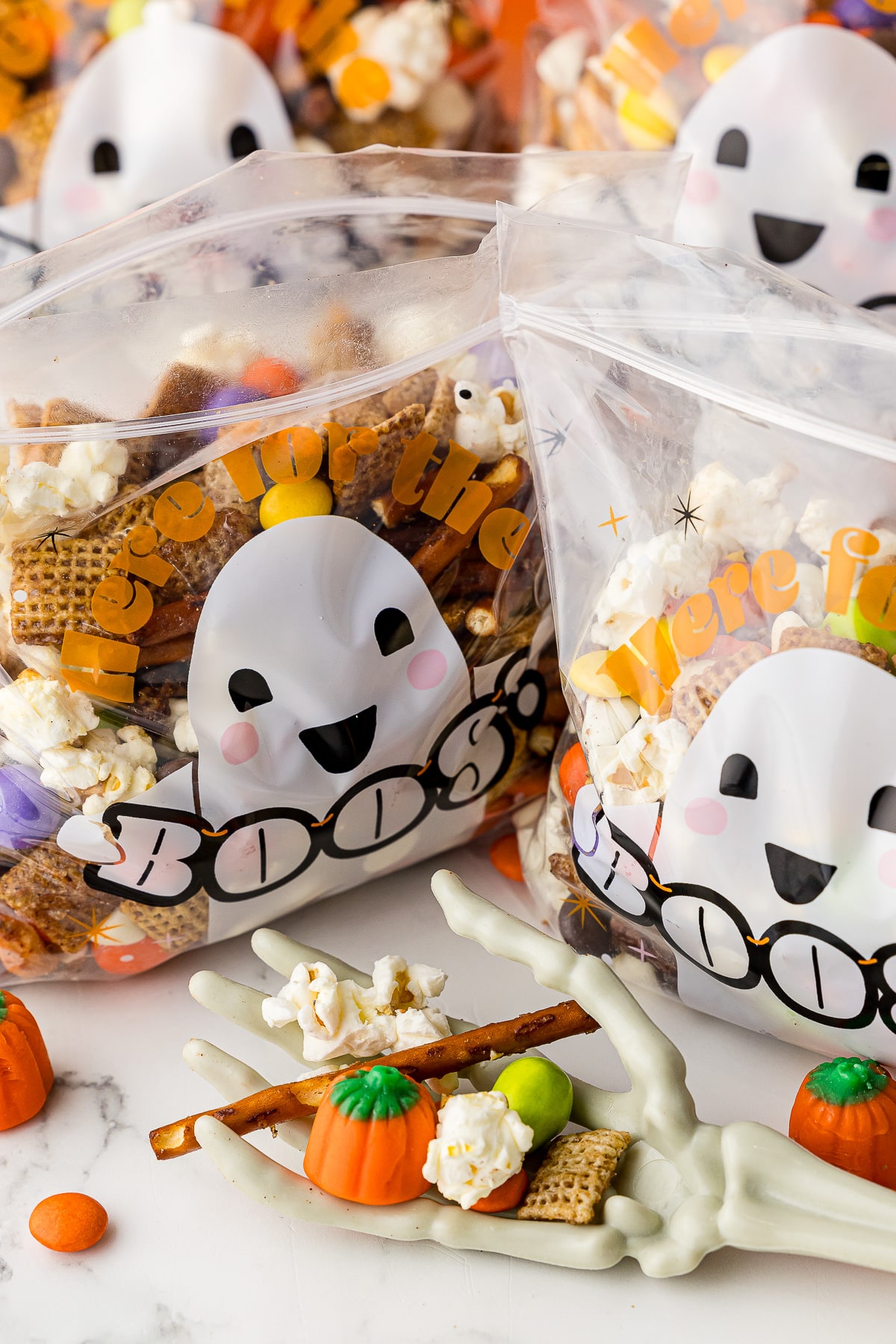 halloween chex mix in treat bags that say where for the boos, a skeleton hand holds a mini pretzel, candy pumpkin, m&ms, and rice chex