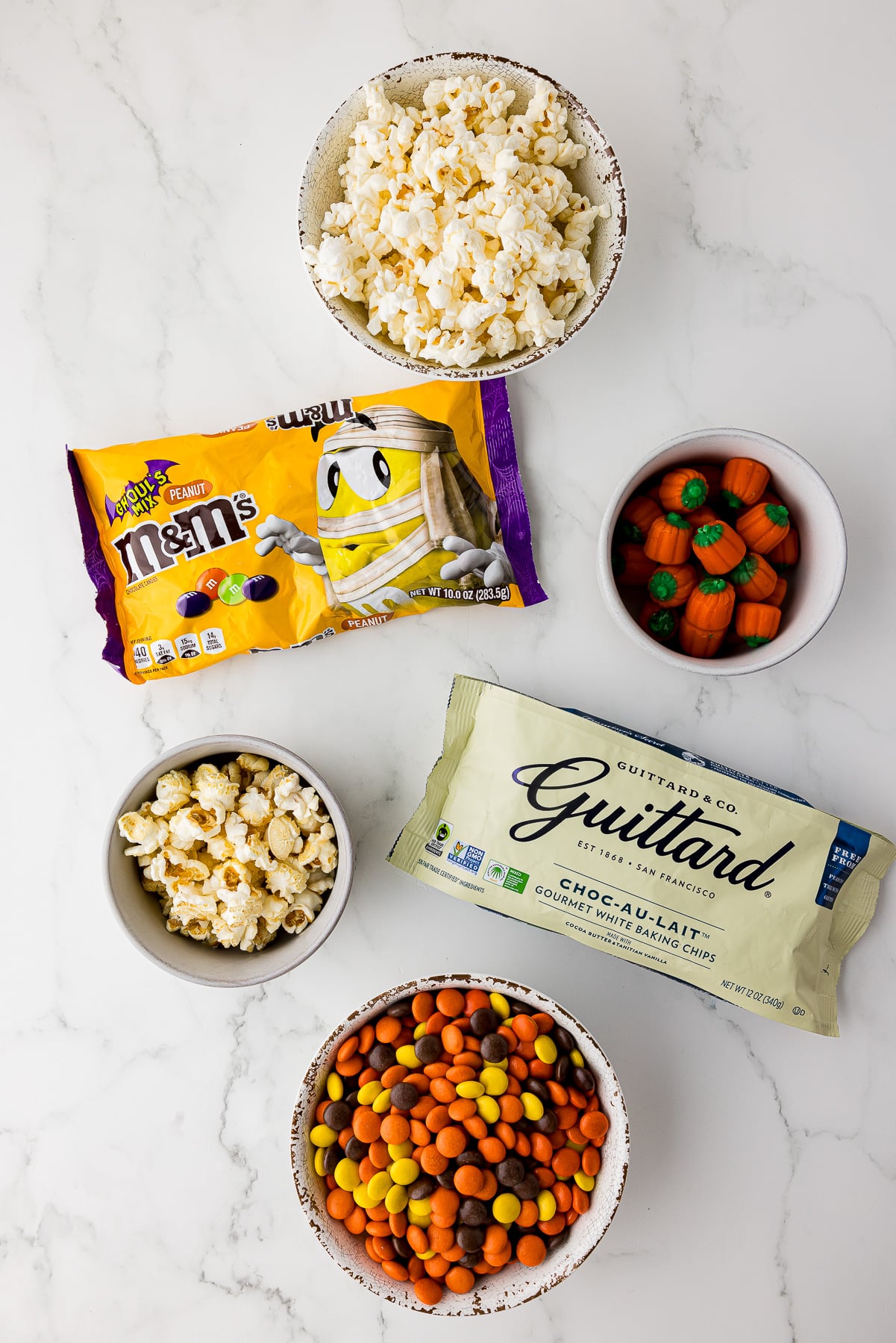 a bowl of popcorn, a bowl of mini candy pumpkins, a bag of ghoul's mix m&ms, a bag of Guittard white chocolate chips, a bowl of reese's pieces, and a bowl of caramel popcorn.