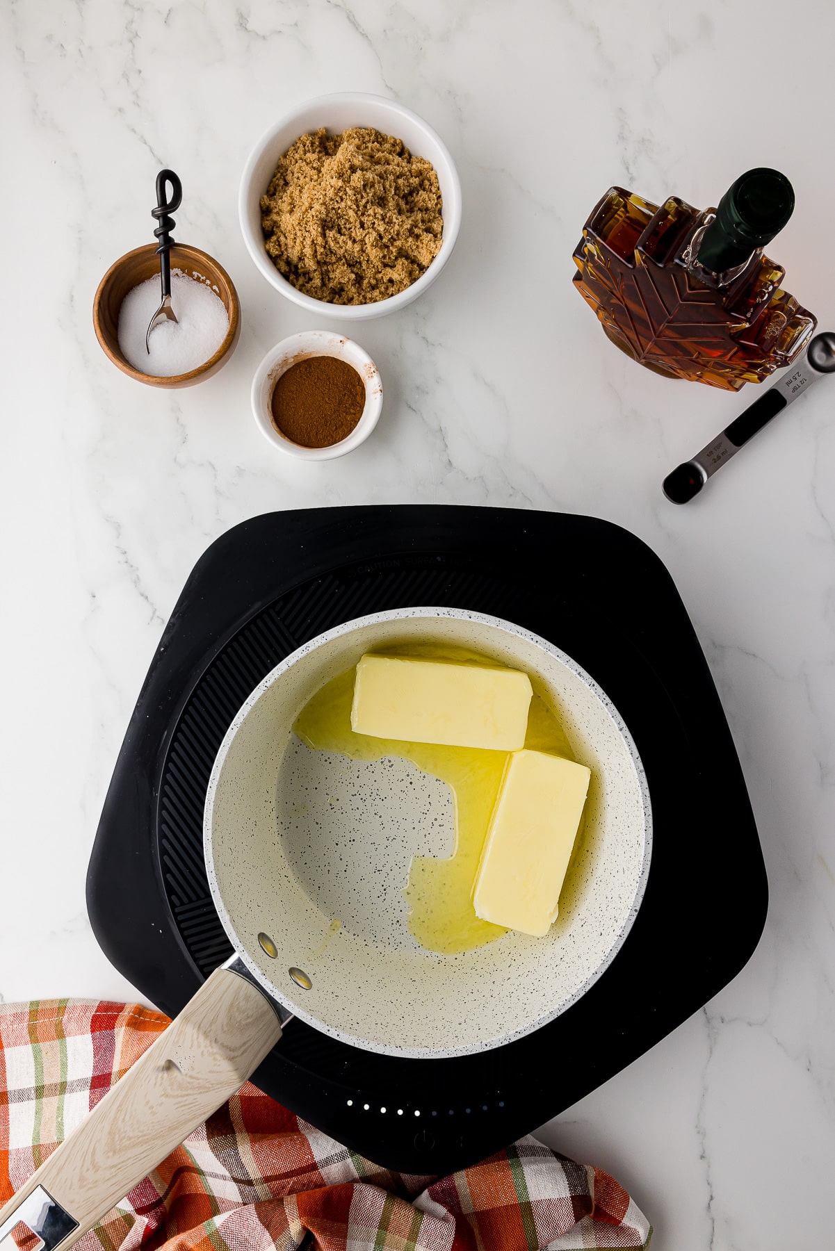 small saucepan with two cubes of butter on a small cooktop on a white marble counter with salt, cinnamon, brown sugar, and maple syrup in the background, with a fall napkin on the counter