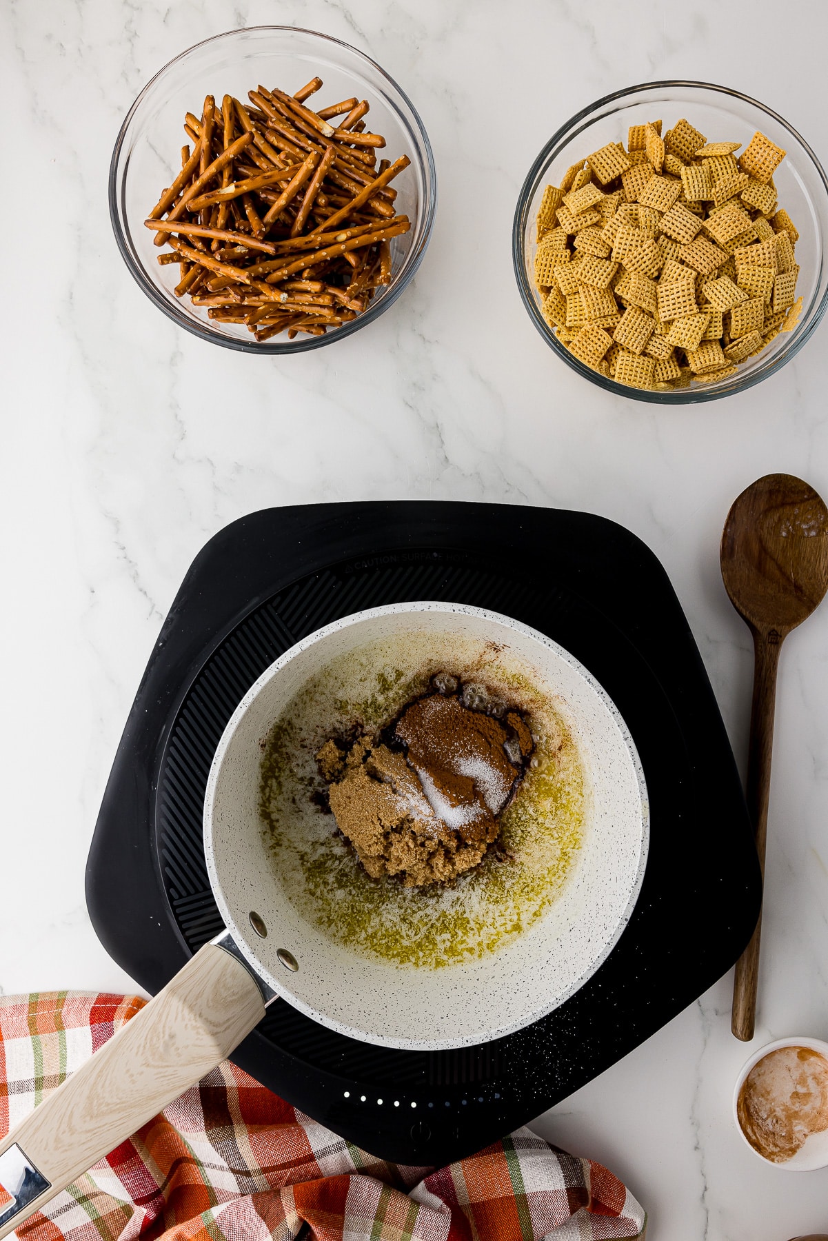 small saucepan with butter, salt, cinnamon, brown sugar, and maple syrup on a small cooktop on a white marble counter with bowls of corn chex and mini pretzles and a wooden spoon.