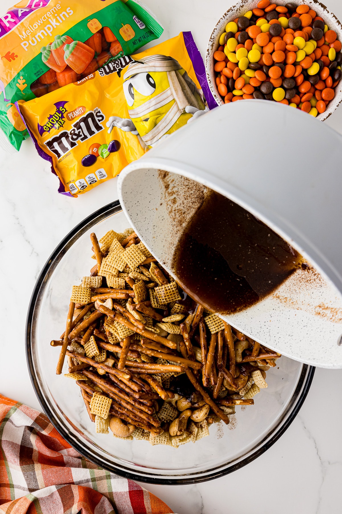 melted sugar coating pouring into a bowl of rice chex, corn chex, mini pretzels in a large glass bowl. Ghoul's mix peanut m&ms, and reese's pieces on the counter.