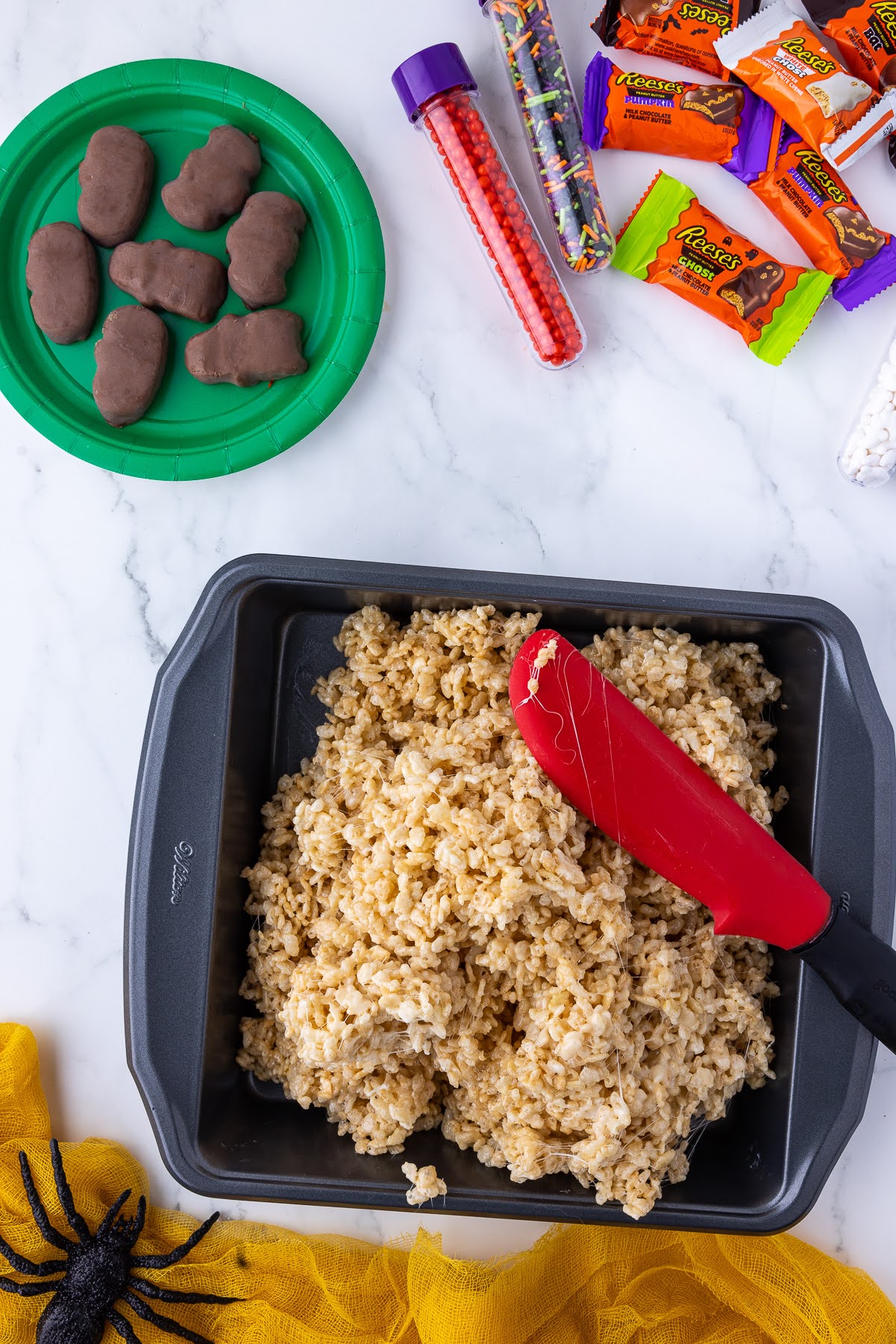 spreading rice crispies into a square wilton pan with wrapped candies in the background and a black spider