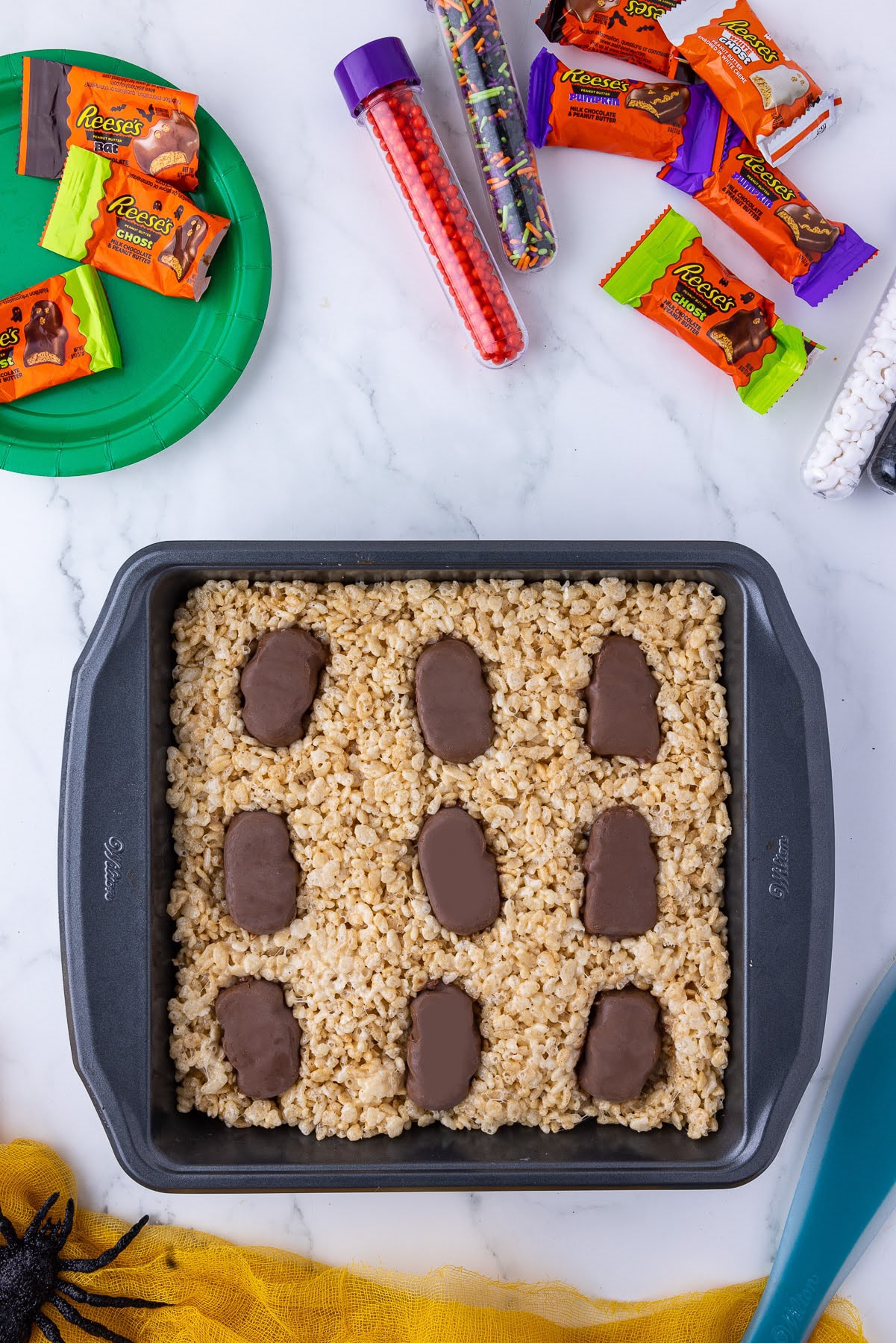 square wilton pan with rice crispie treats with reese's wrapped candies and halloween sprinkles and wilton sprinkes in plastic viles