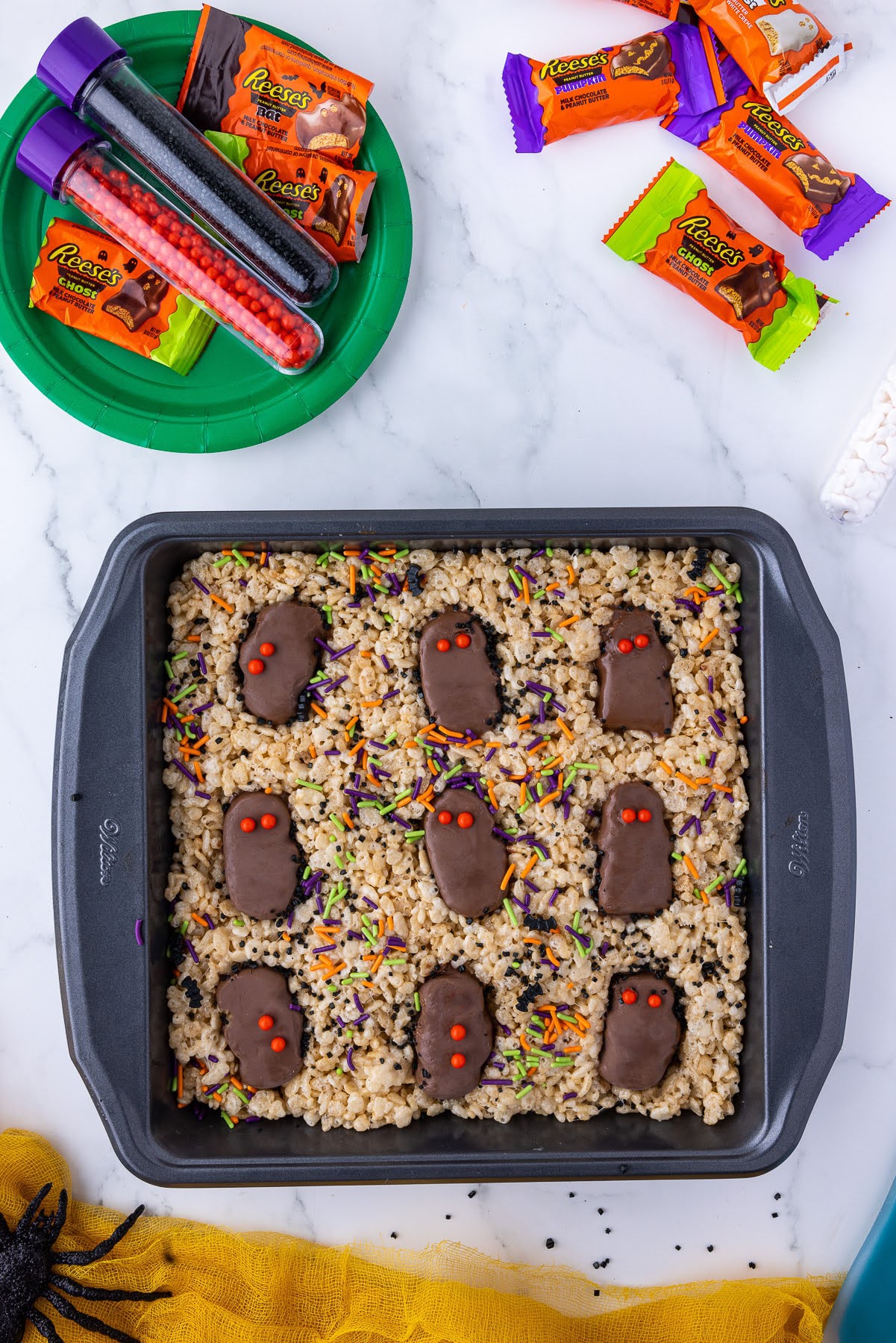 square wilton pan with rice crispie treats with reese's wrapped candies and halloween sprinkles and 2 orange eyes on each one