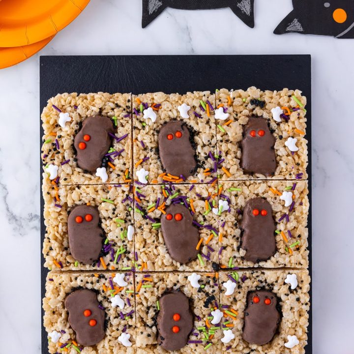 six rice crispie treats on a black slab with reese's peanut butter cups, wrapped candies, orange paper plates and bat napkins
