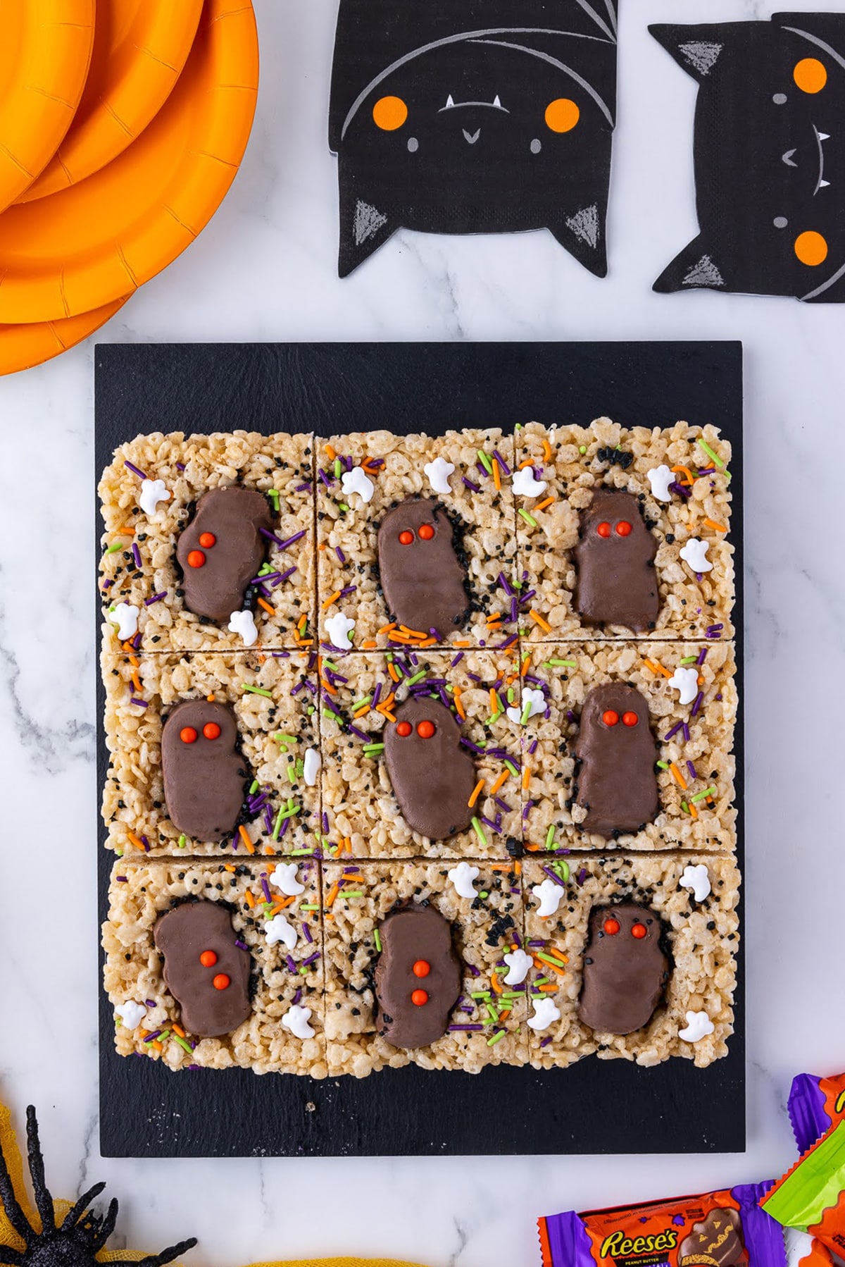 six rice crispie treats on a black slab with reese's peanut butter cups, wrapped candies, orange paper plates and bat napkins