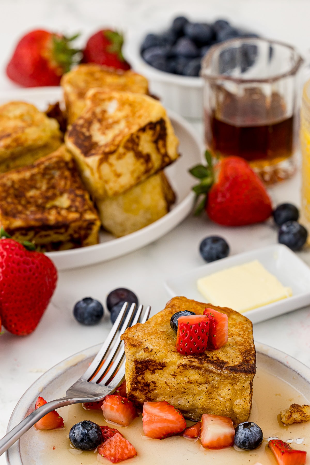 plate of french toast with strawberries, syrup, and blueberries, plaid cloth, a fork, strawberries, blueberries, and syrup in the background with pats of butter