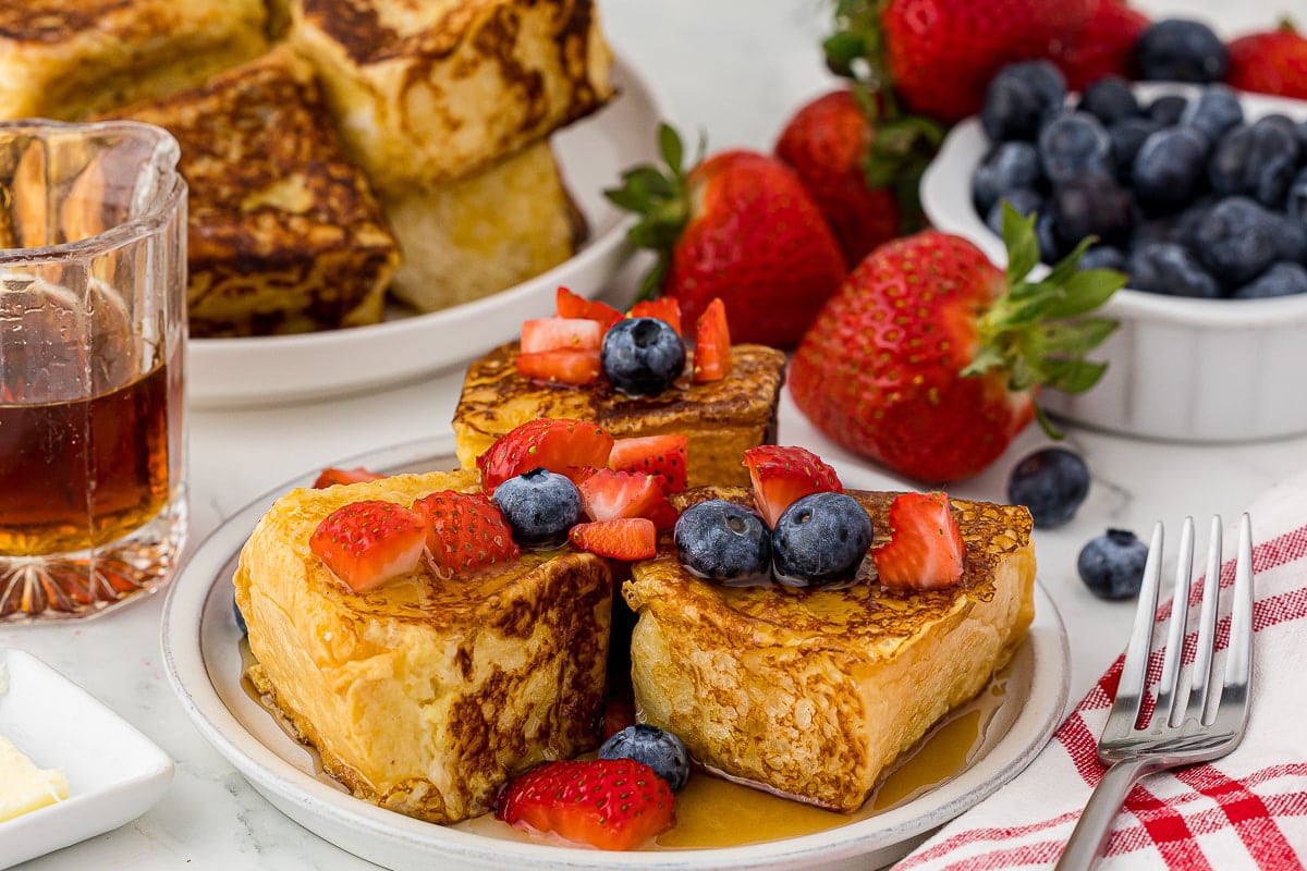 plate of french toast with strawberries, syrup, and blueberries, plaid cloth, a fork, strawberries, blueberries, and syrup in the background
