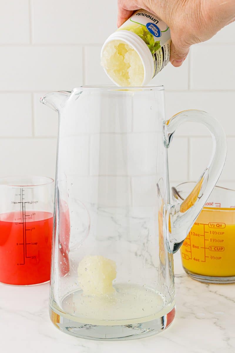 limeade pouring into glass pitcher with orange juice and grapefruit juice in the background