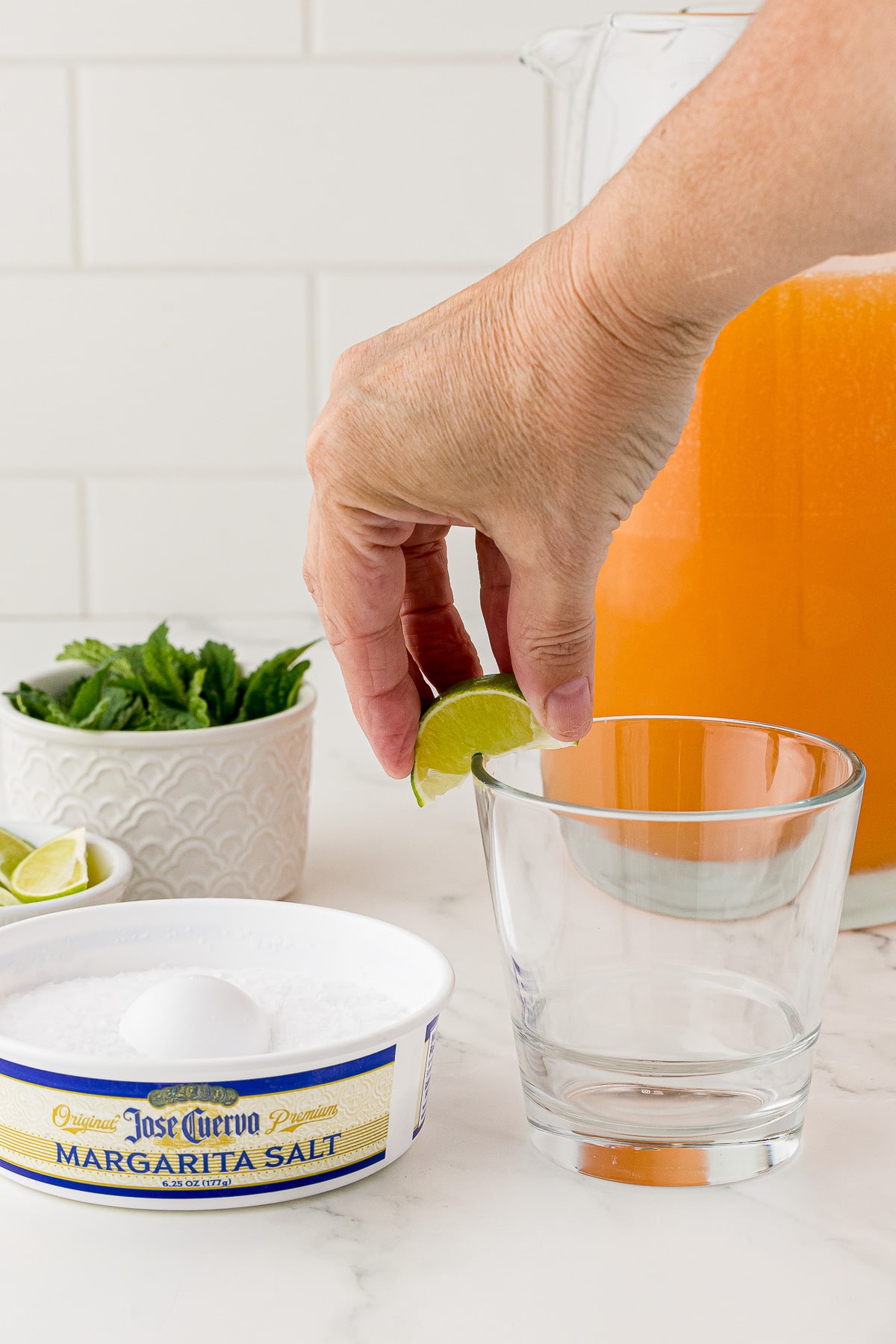 rimming a glass with a lime, margarita salt, mint leaves, and a pitcher of the mixture in the back