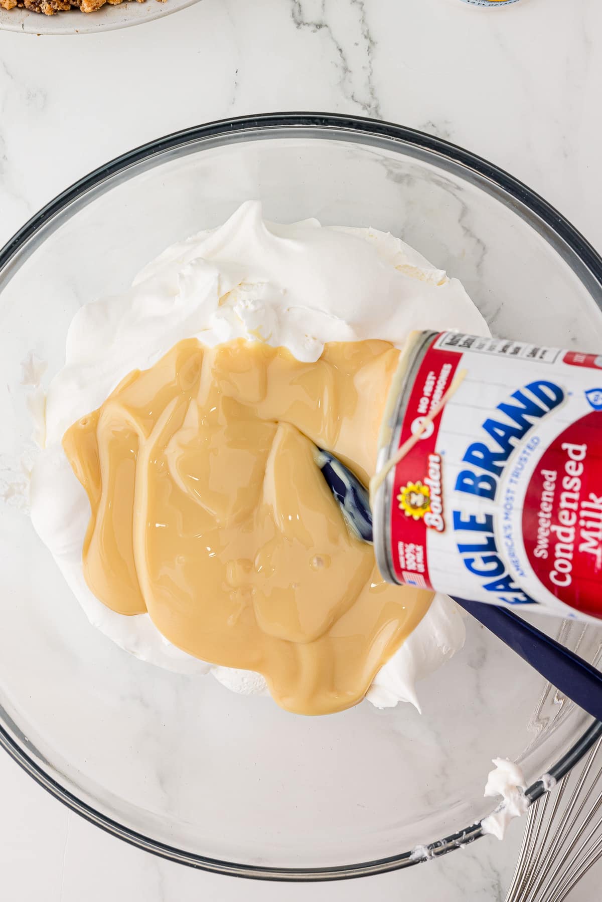 Pouring condensed milk onto cream cheese in a clear glass mixing bowl