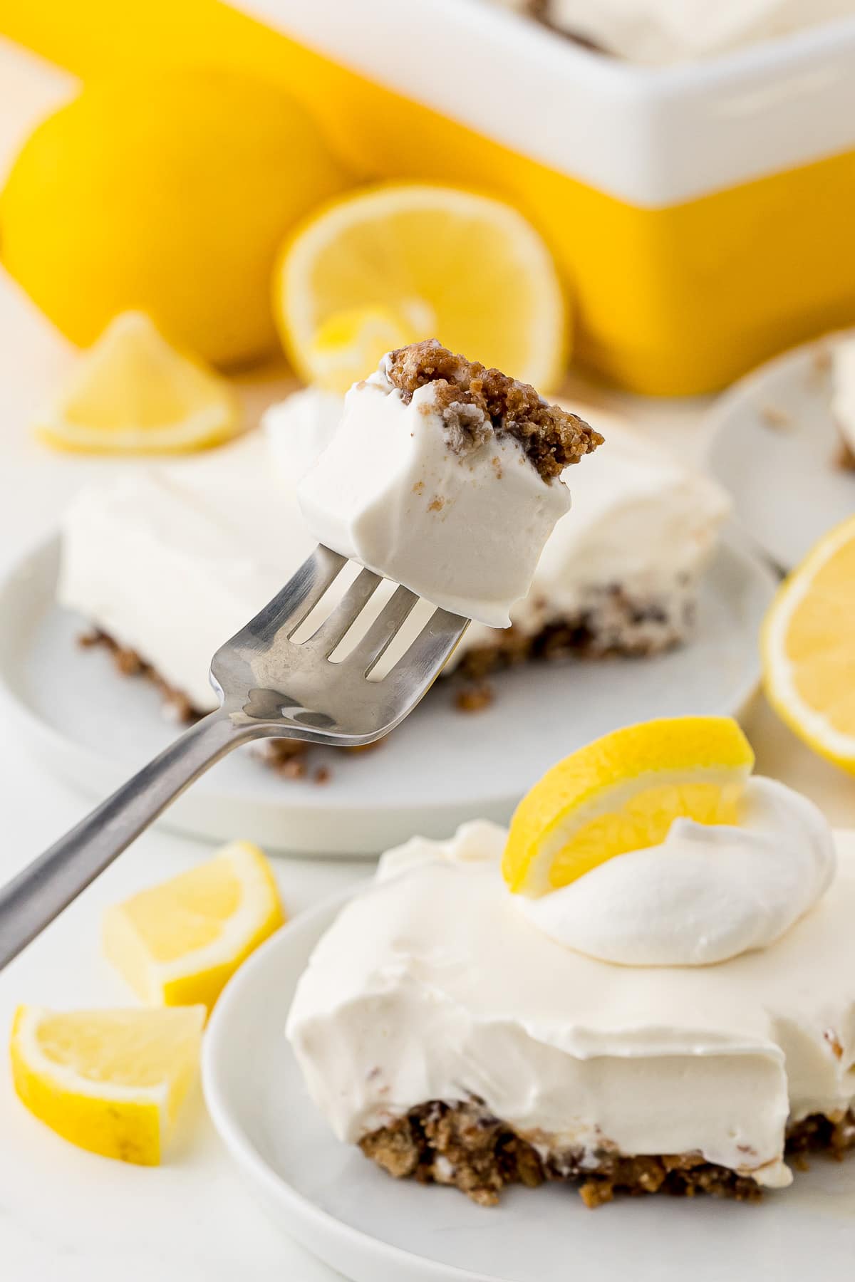 A fork holding a bite of lemon pie with additional plates of pie and lemons in the background