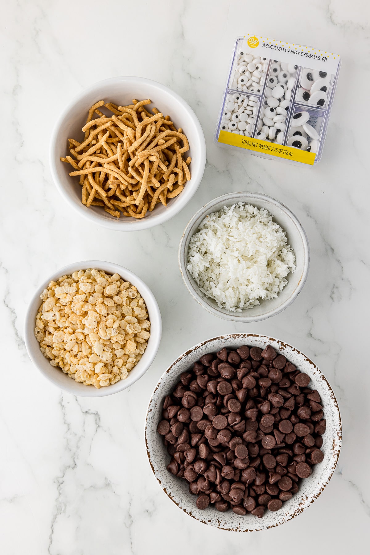 four white bowls filled with chocolate chips, rice krispie cereal, shredded coconut, chow mien noodles, and a box of wilton candy eyes