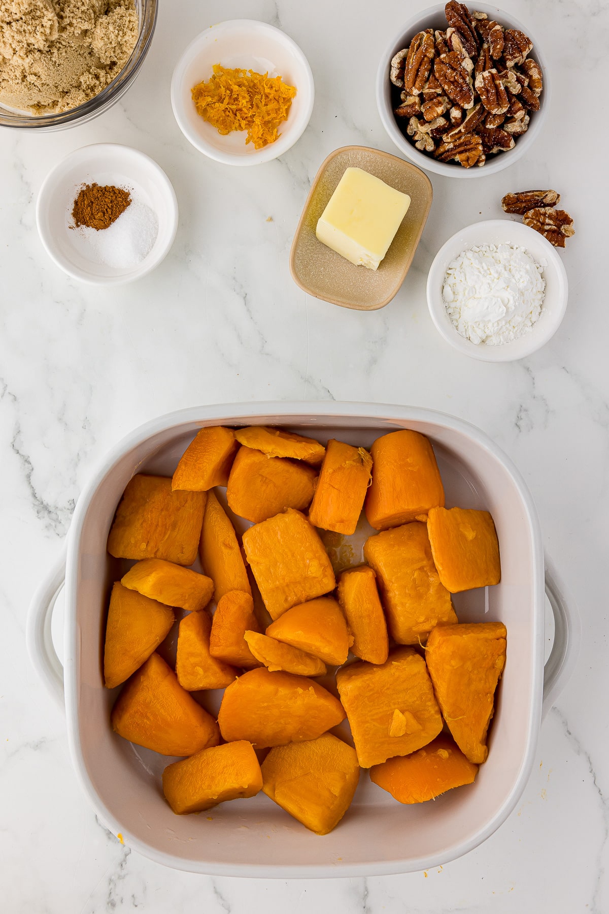 plain sweet potatoes in a shite square casserole dish with small bowls filled with brown sugar, cinnamon, salt, orange zest, butter, pecans, and cornstarch