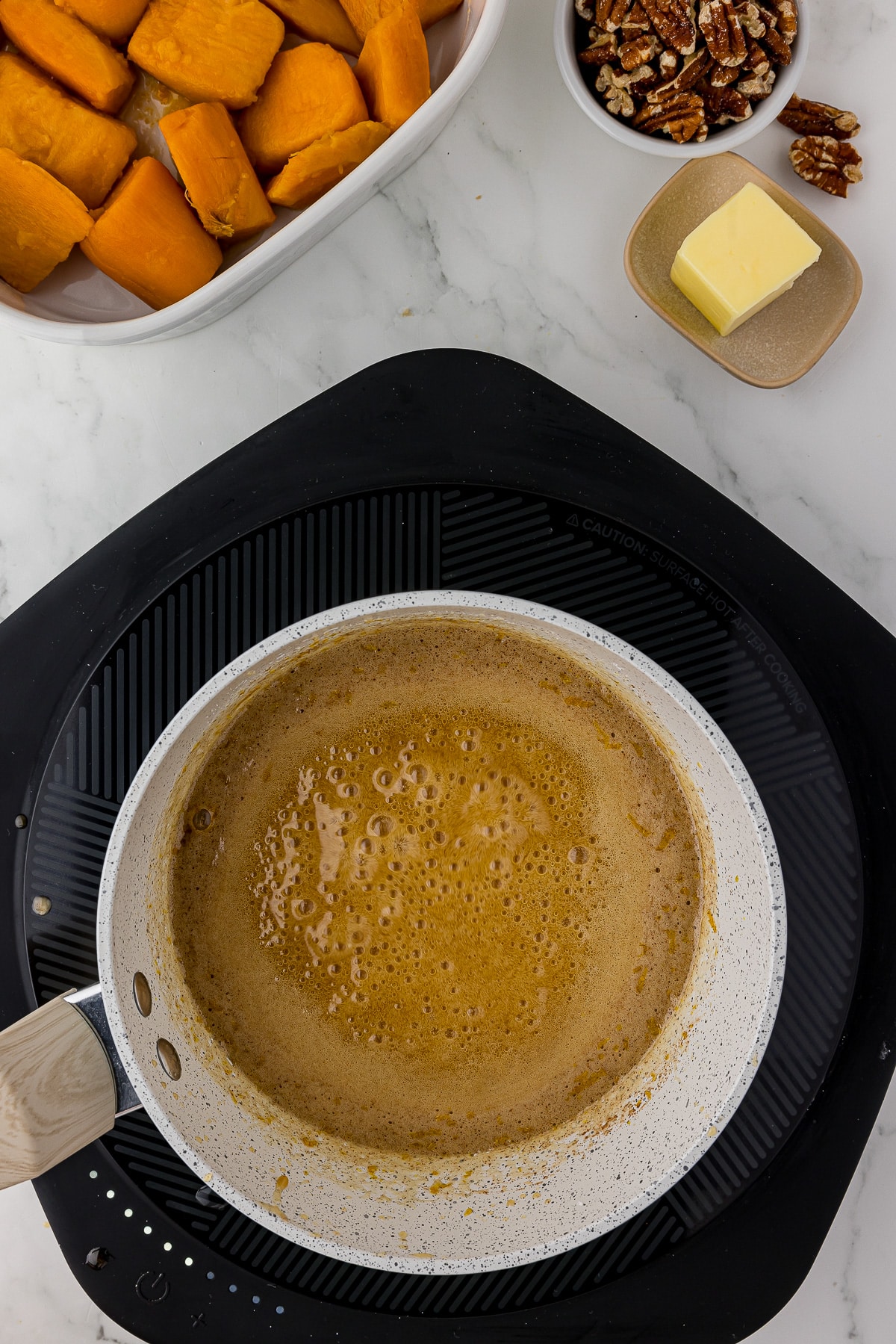 bubbling brown sugar mixture in a saucepan on a heating plate with sweet potatoes, pecans, and butter on the counter