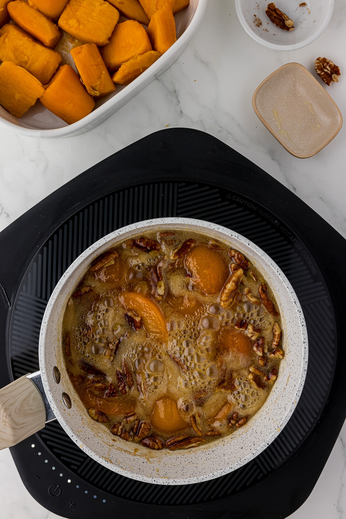 Boiling brown sugar mixture with apricots, butter and pecans in a saucepan on a heating plate on a white marble countertop