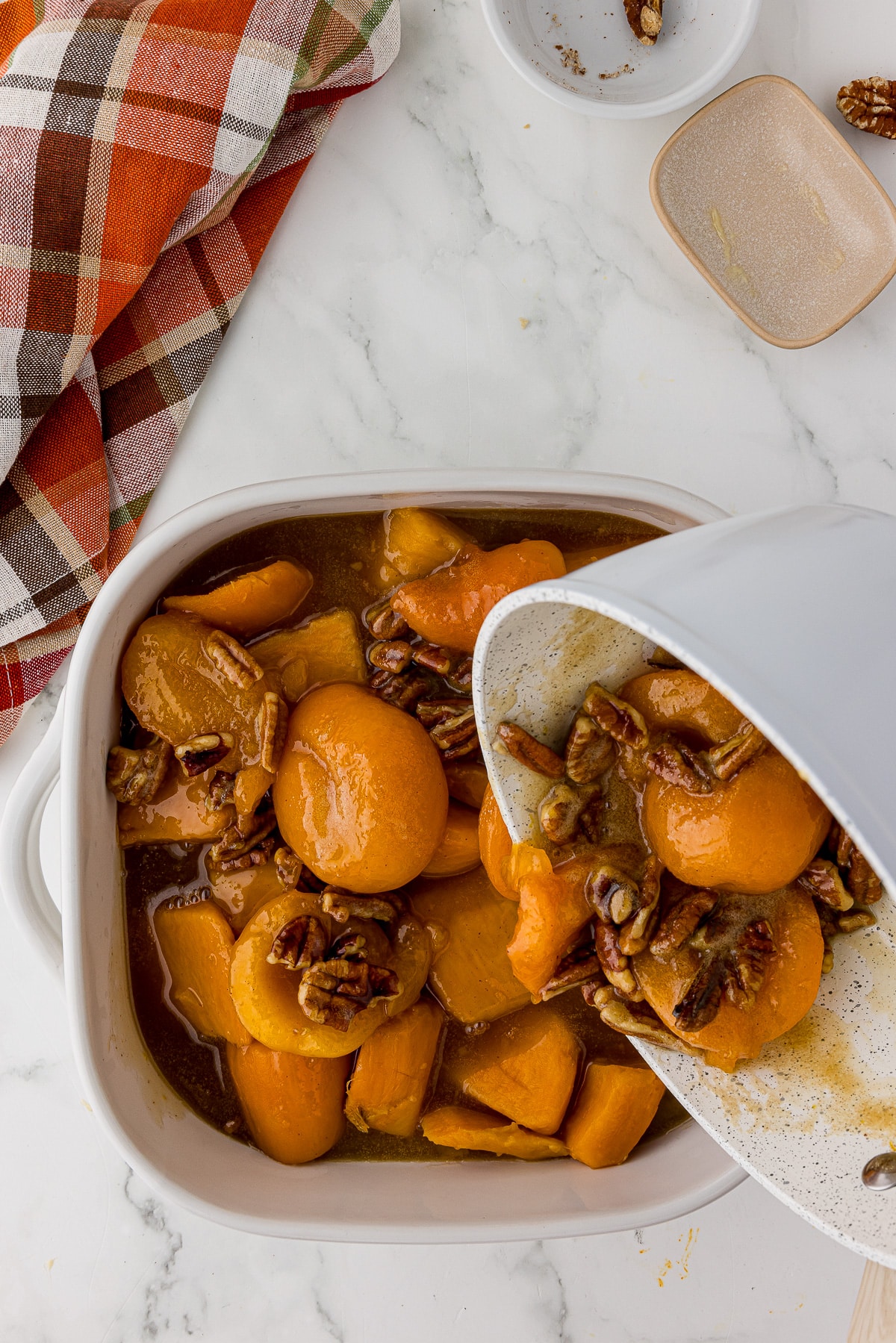 white casserole dish with sweet potatoes and the brown sugar mixture being poured over with an autumn napkin on a white countertop