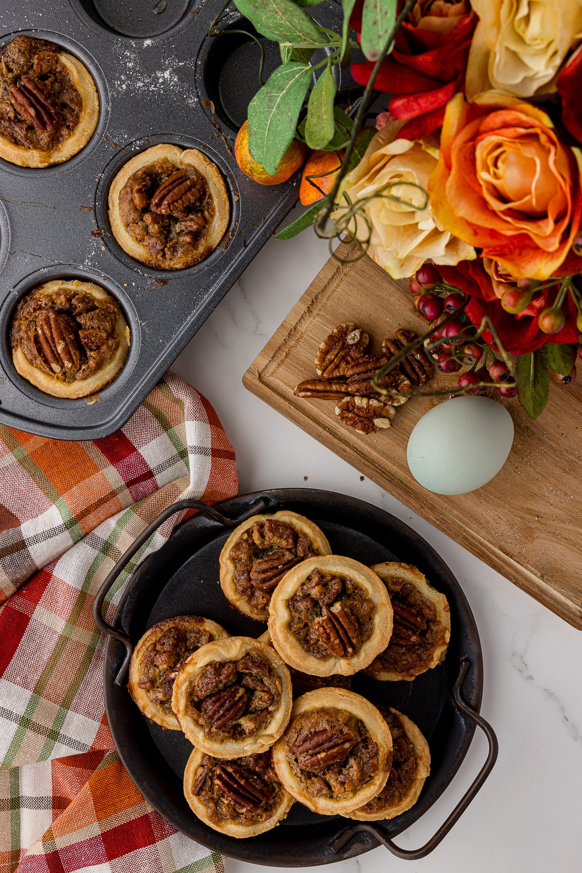 Mini pecan pies stacked on a metal plate with a mini muffin pan, a plaid autumn napkin and a cutting board.